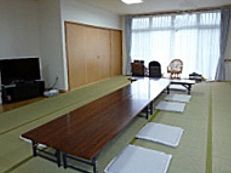 広い畳敷きの部屋。突き当りに掃き出し窓がある。窓と直角に長テーブルがあり、座布団が並んでいる。板の間にテレビが置いてある。