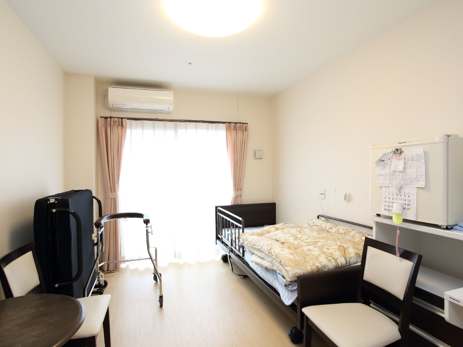 室内は広く明るい。ご入居者様のベッド、折り畳みベッド、歩行器は余裕をもって配置されている。テーブル、いすもあるので来客があっても安心。