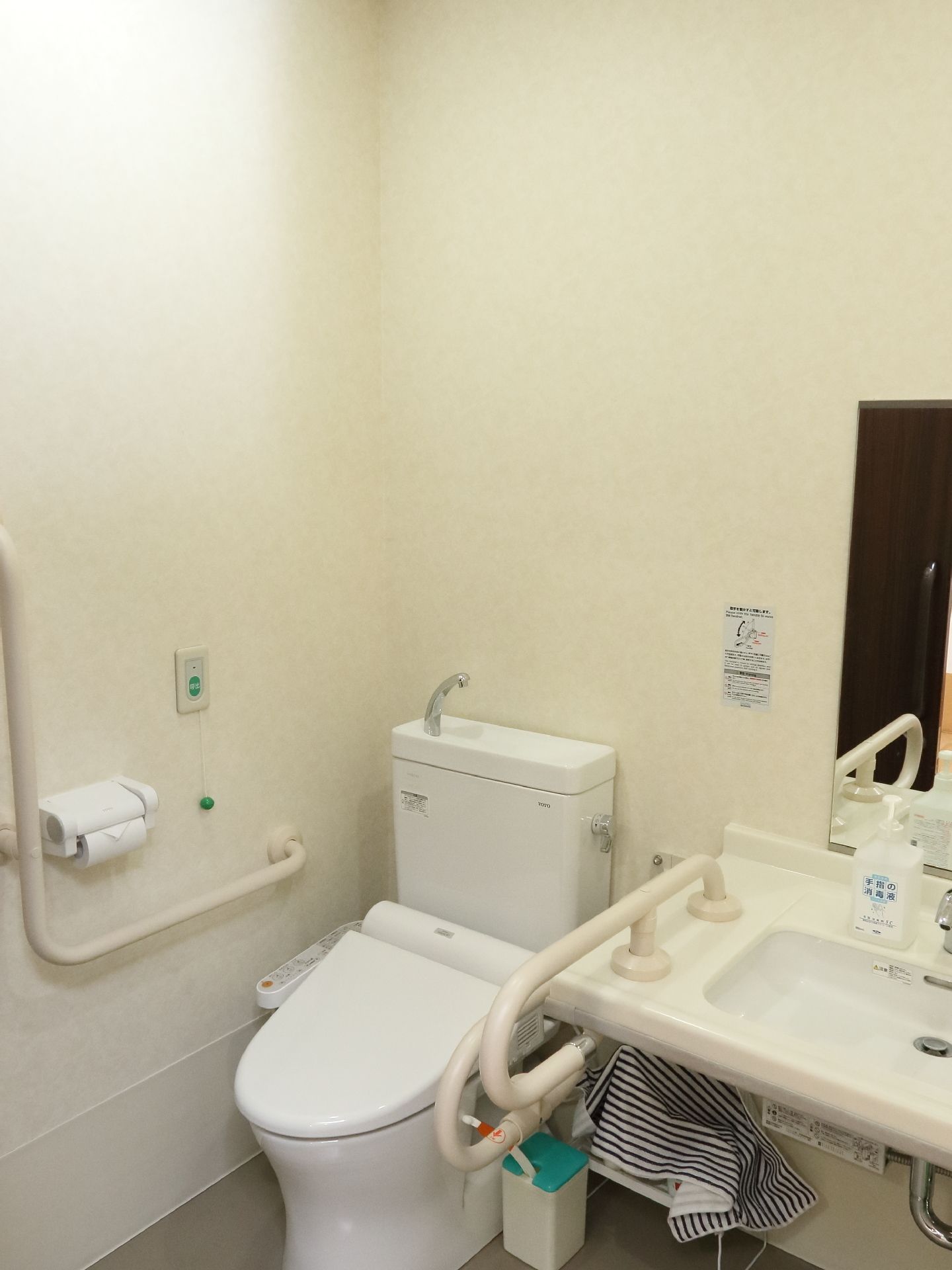 洗面台、左側に手すり設置。トイレはウォシュレット付きで、ヘルプコールボタンが設置。トイレ左側にはエル字型の手すりが設置されている。