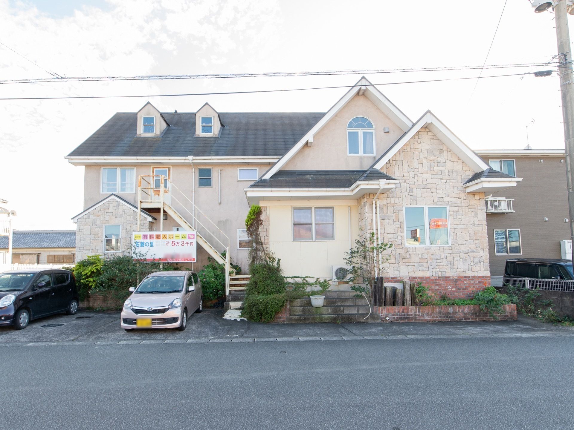 ベージュの外壁で三角の屋根になっており、おしゃれな雰囲気がある。建物の左側には看板が立っており、前には車が停まっている。