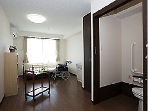居室はワンルーム。手前右の引き戸が開いており中はトイレになっている。奥の部屋にはキャビネットなどがあり壁際にベッドが置かれている。