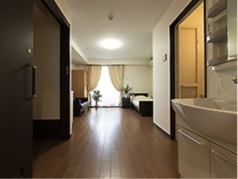 手前に洗面台。広い通路の両側に大きな引き戸がある。奥は寝室になっており右壁際にベッドが見える。そのほかに観葉植物、ソファなども置かれている。