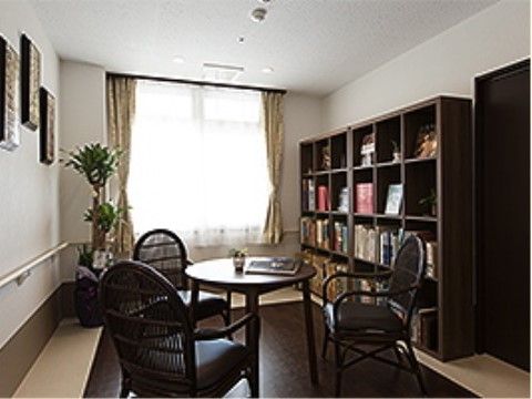 奥の大きな窓から光が差し込む部屋。右側の壁に本棚があり、本や置物がディスプレイされている。手前にはテーブルと椅子が３脚用意されている。