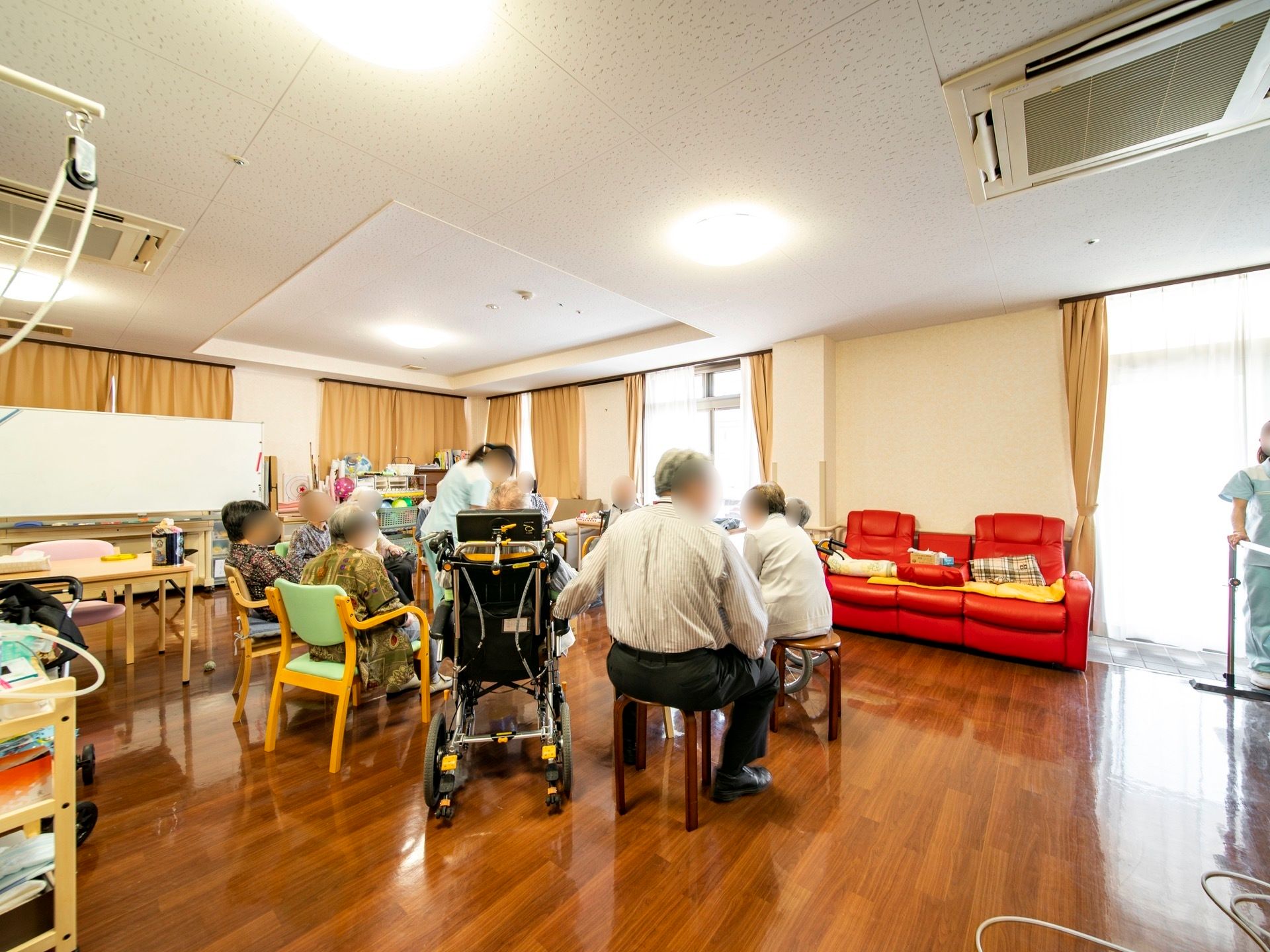 窓際に赤いソファが置かれている談話室がある。ダークブラウンの床の広い空間には、スタッフと入居者様が円になって歓談をしている。