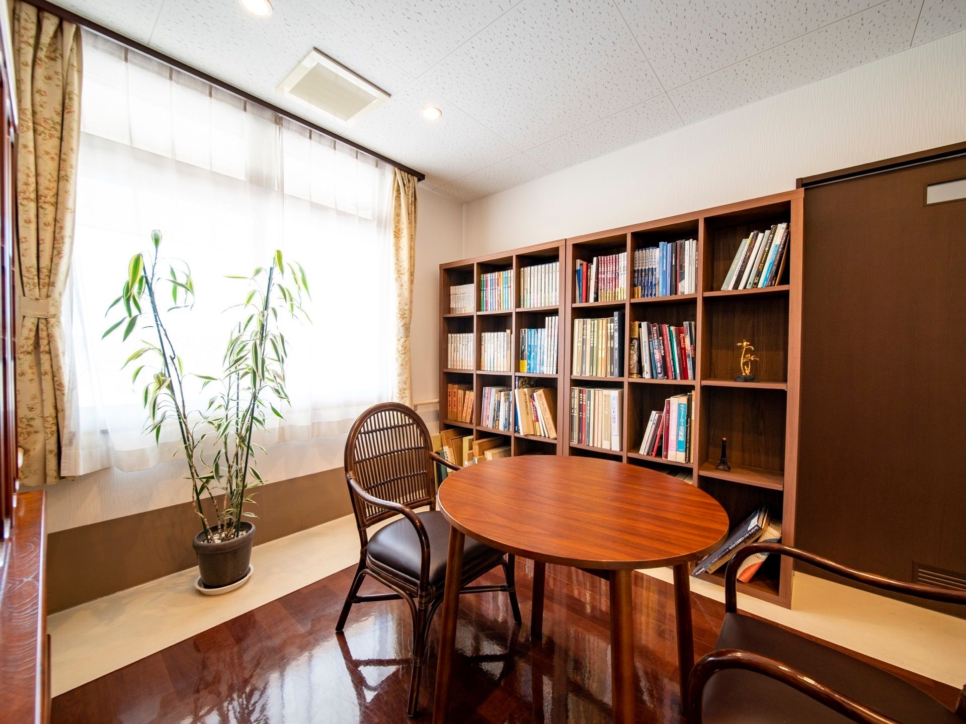 大きな本棚が置かれている小部屋がある。窓にはレースのカーテンが掛かっている。テーブルと椅子が置かれ、大きな観葉植物が飾られている。