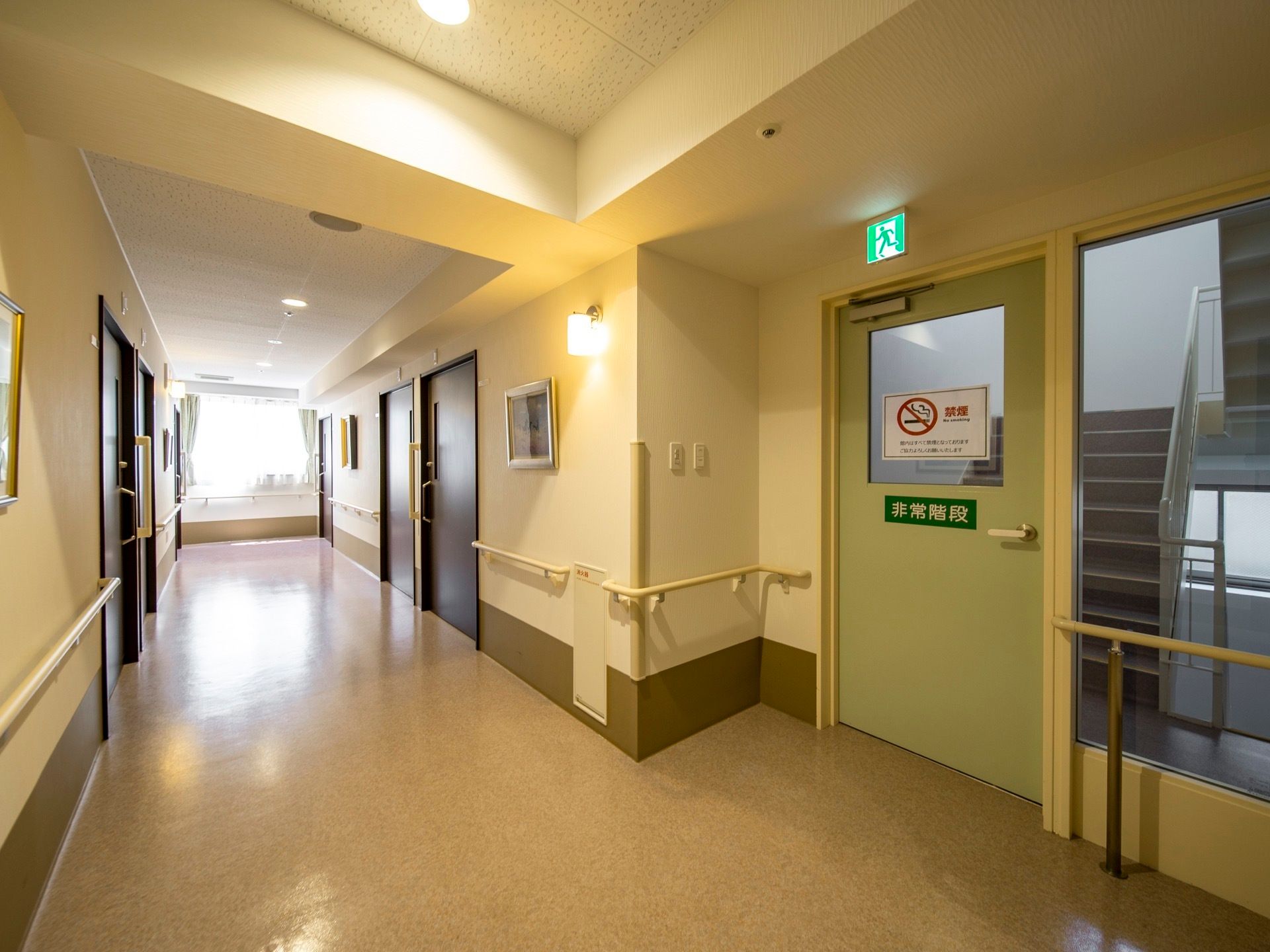 クリーム色の壁の広い廊下がある。入居者様の居室のドアが並び、壁には手すりがついている。非常階段があり、壁にはガラス窓がはまっている。