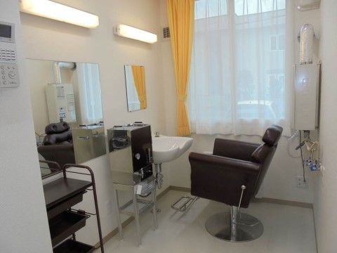 窓がある個室に設けられた理美容室である。白色の壁には鏡が備わり、洗髪台の前には茶色の椅子が置かれている。