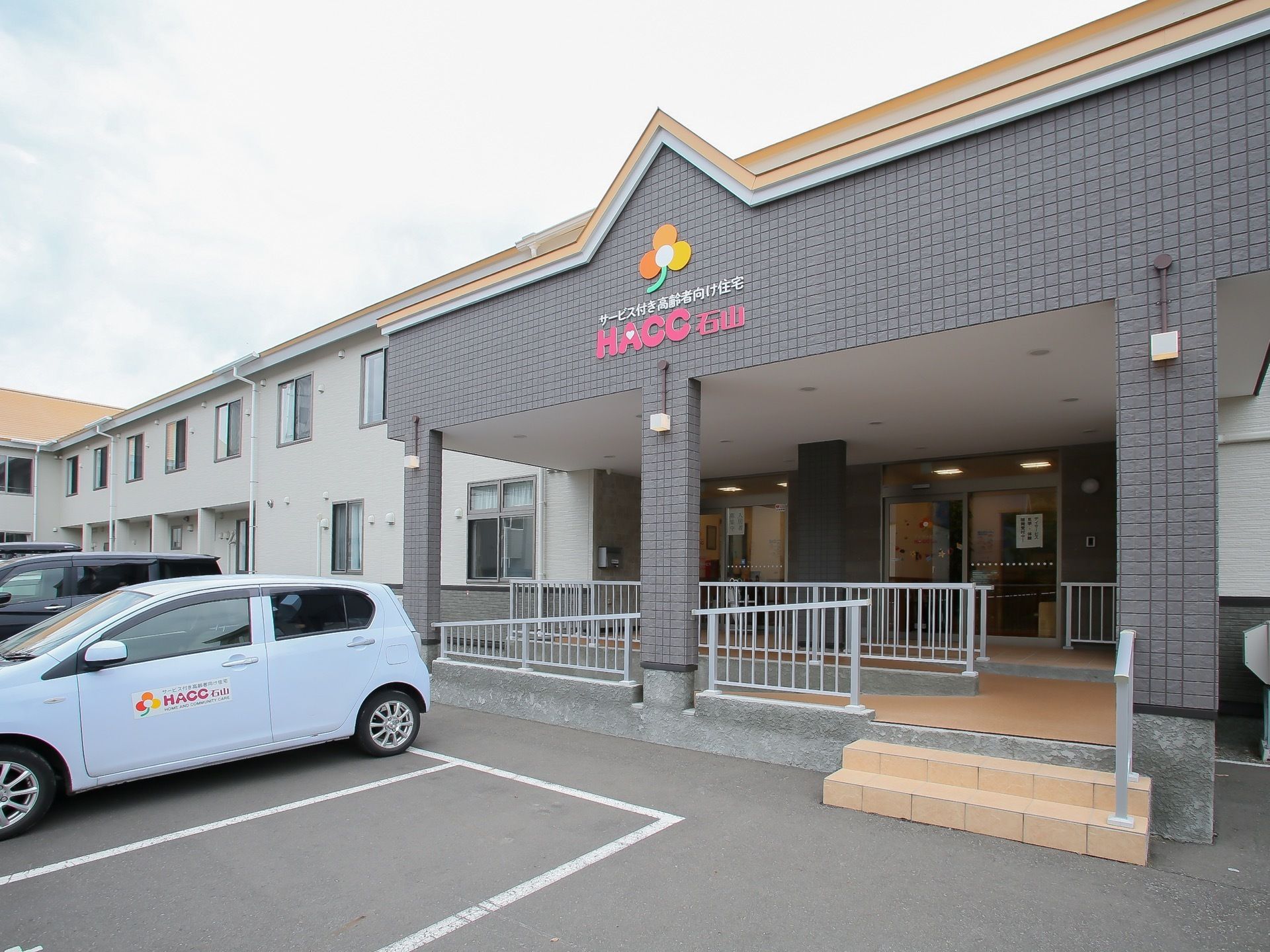 グレーの外壁が印象的な、広々としたエントランスである。手すりが備えつけられている。建物の目の前には、駐車場も完備されている。
