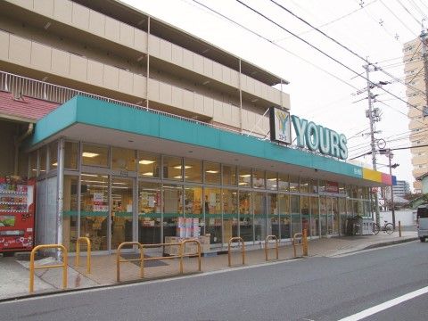 近く建つスーパーマーケット。屋根は緑色で、外観は全面ガラス張りになっている。隣に自動販売機が設置されている。