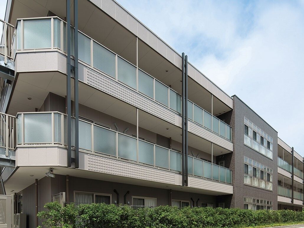 ３階建ての横に広い建物で、１階の前には背の低い木が植えられており、２階と３階にはベランダが備わっている。