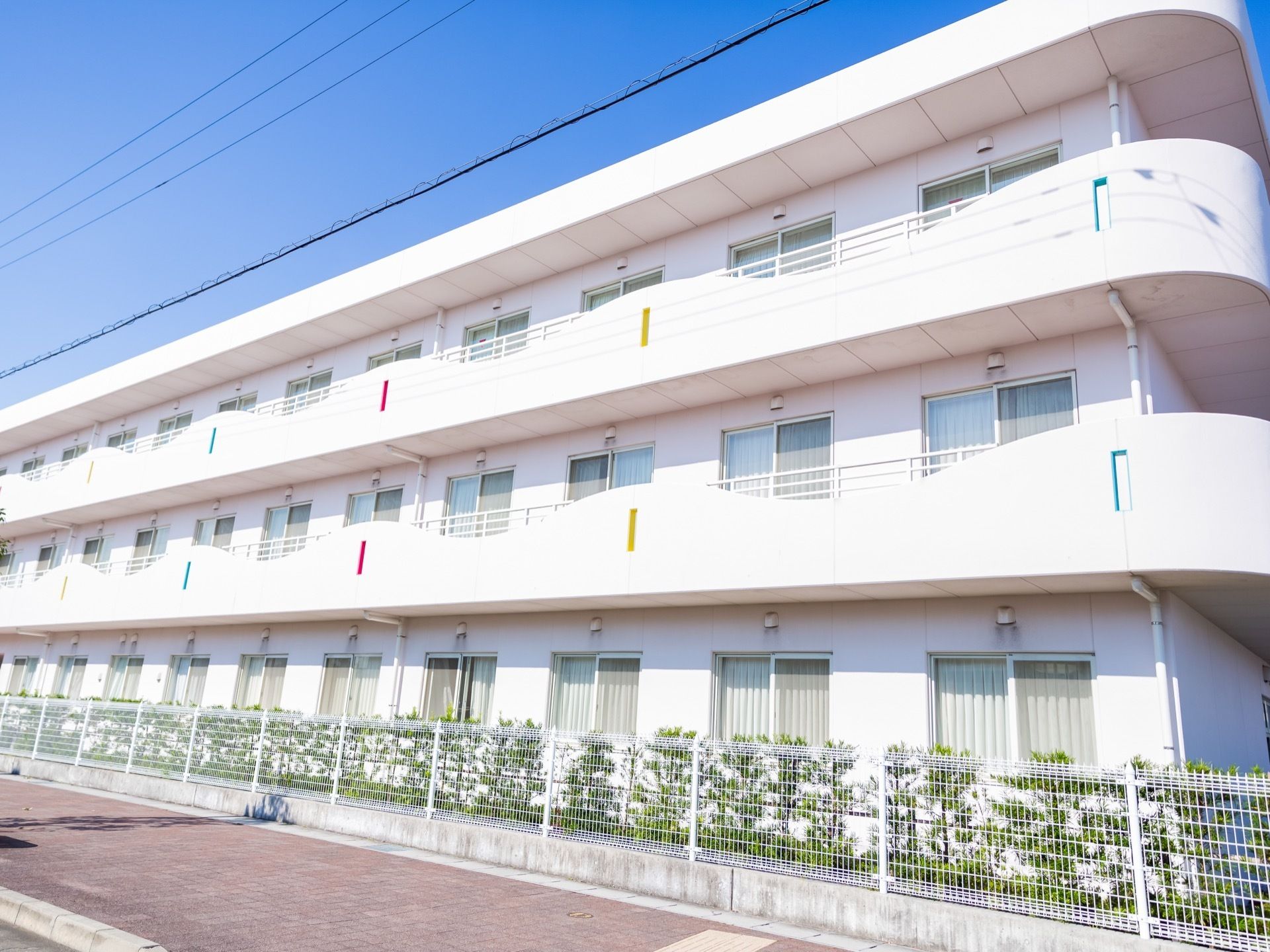 アイボリーホワイトの外壁で統一された３階建ての施設である。２階と３階の居室の窓の外には柵のついたベランダがある。