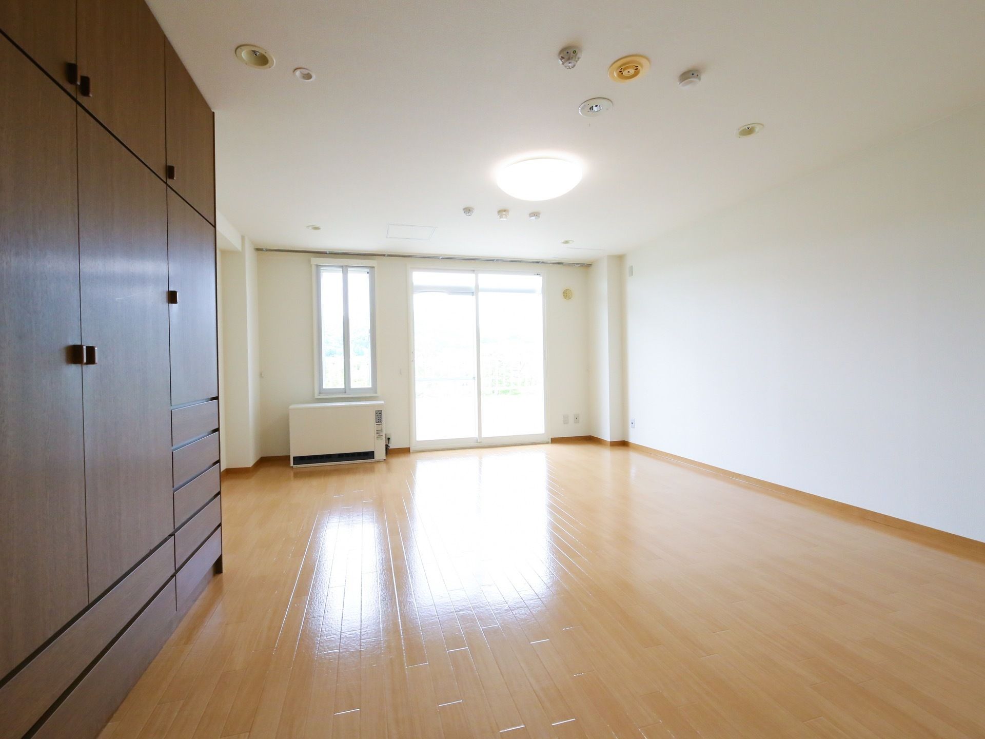 部屋の奥の壁面には、掃き出しの窓と細長い窓がある。左手前には、大きな木製の収納家具が設置されている。