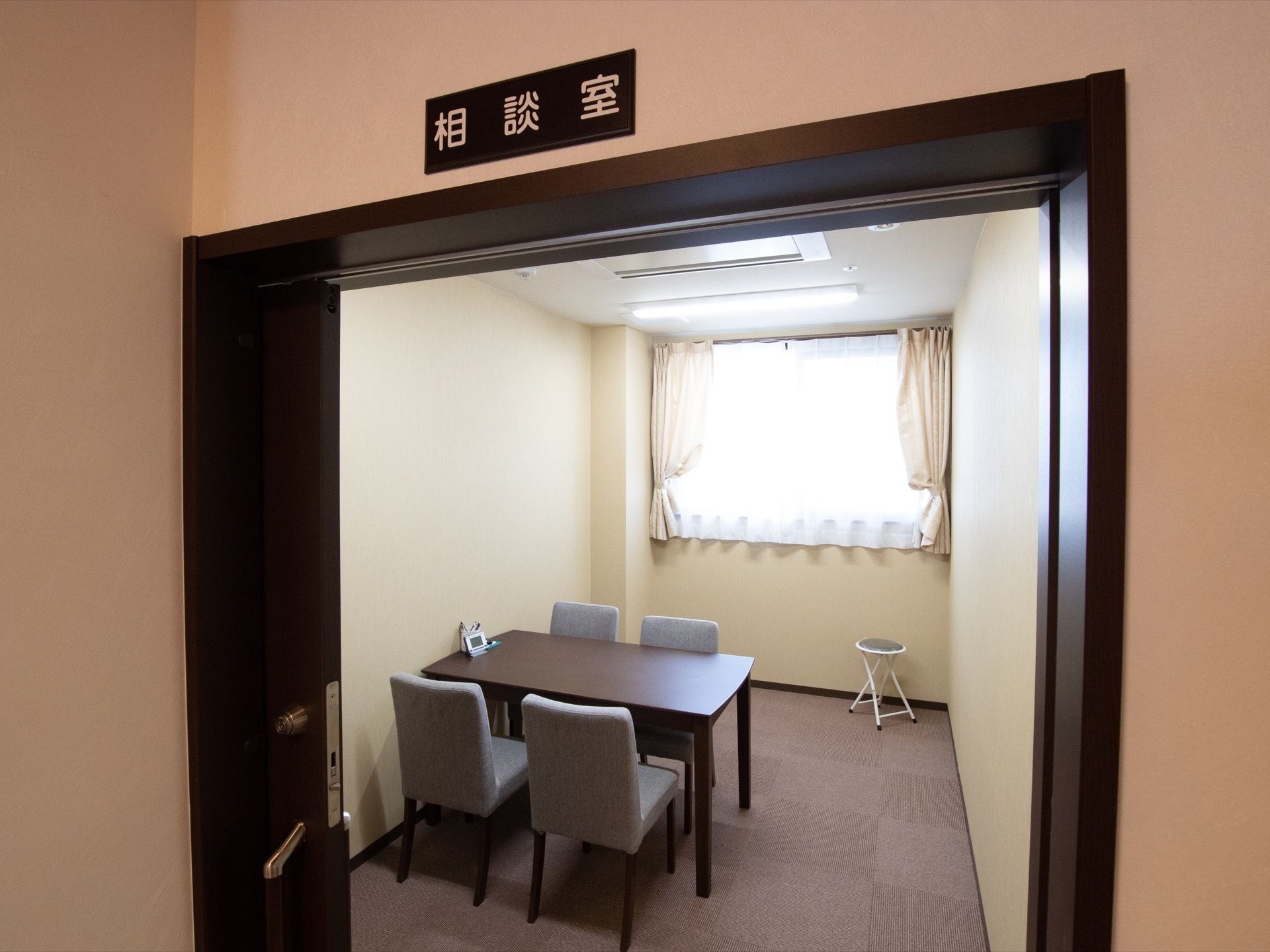 相談室と表示されたプレートが取り付けられている。木製のテーブルのまわりに椅子が４脚置かれている。窓にはカーテンがかかっている。