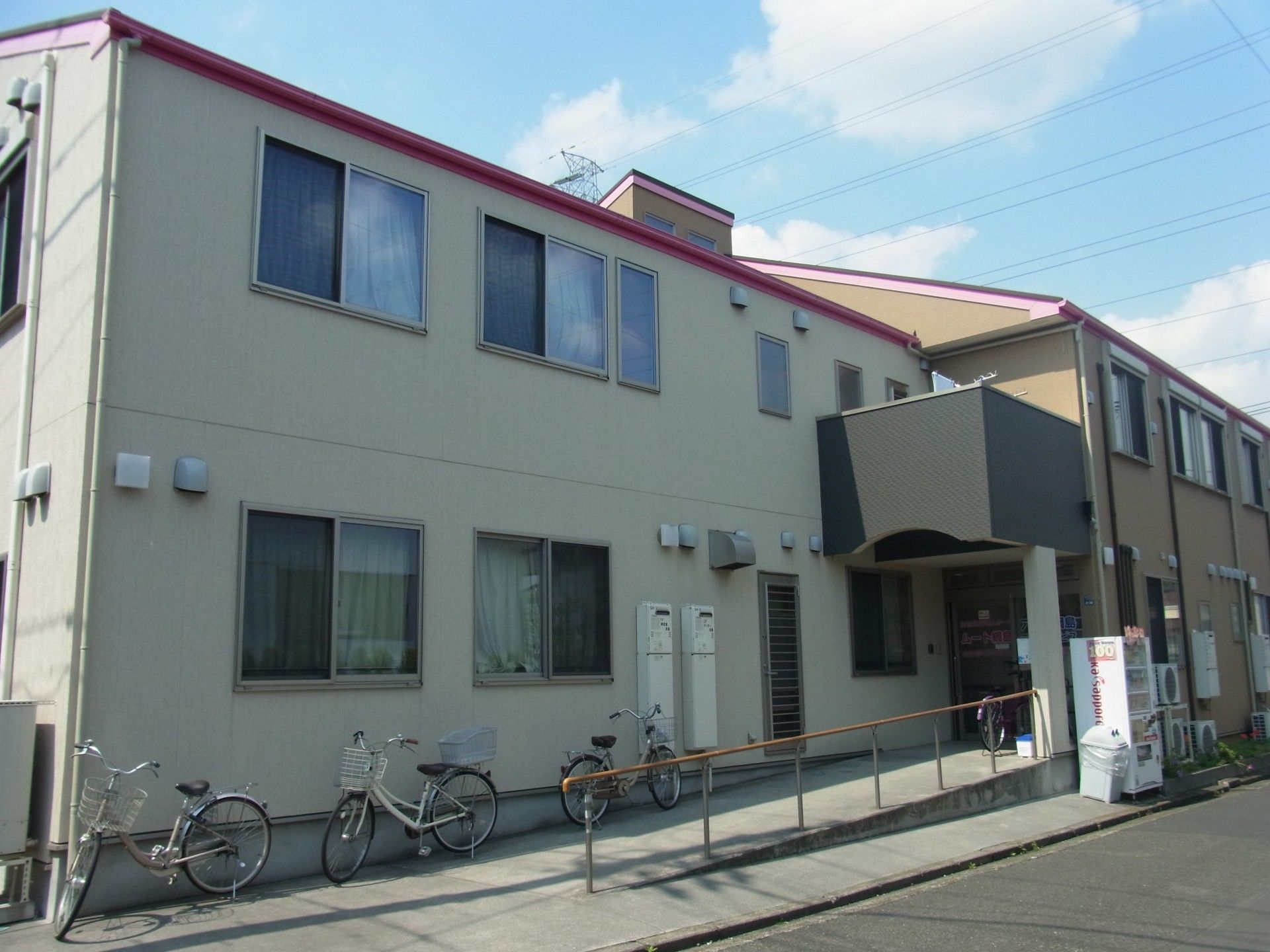 ２階建ての施設の建物である。玄関の前に続くスロープがあり、手すりが備わっている。内覧会の案内の看板が置かれている。