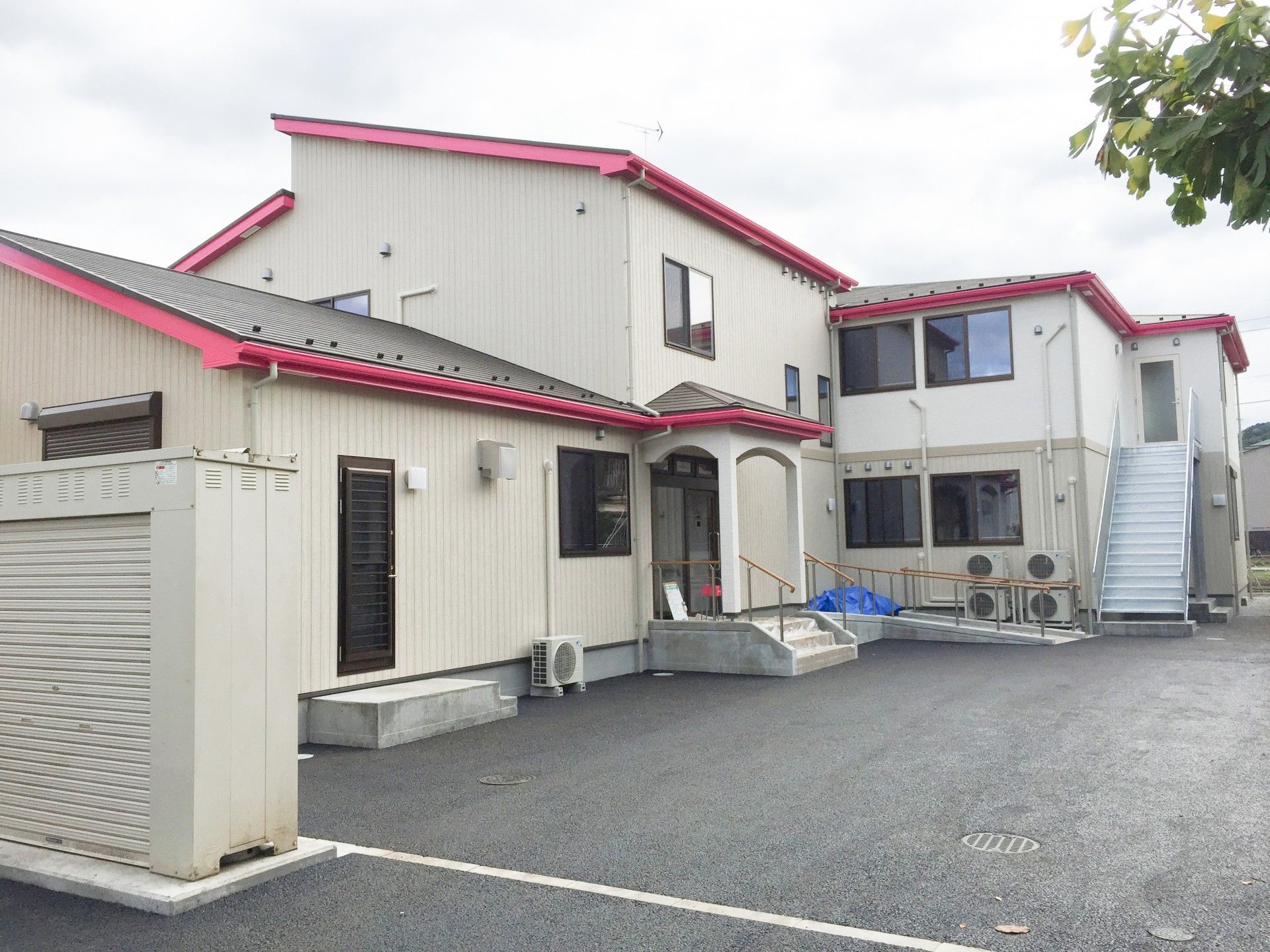 赤い屋根を持つホームは２階建てである。道路からの通路や建物周りの植え込みを整備して、入居者様や来訪者様を迎え入れている。