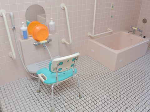 淡いピンクを基調とした広い浴室。手すりなどの安全対策が取られ、椅子や洗面器などの小物は目立ちやすいビビットな色で揃えている。