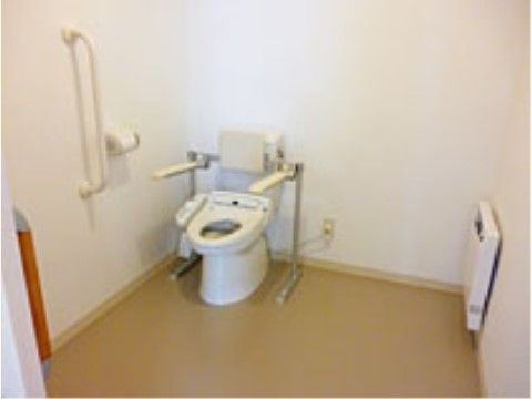 トイレ内のスペースは広く、温水洗浄便座が備えられている。便座には背もたれ、手すりが設置されており、床と壁は明るい色で統一されている。