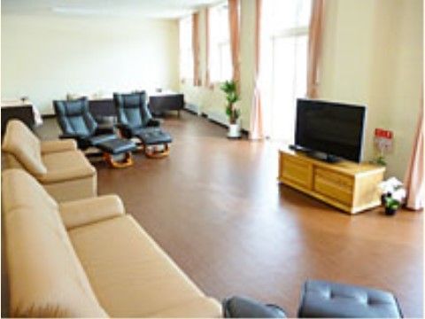 広いスペースに大きな窓があり、観葉植物が置かれている。室内は明るく、大型テレビの周りにソファが設置されている。
