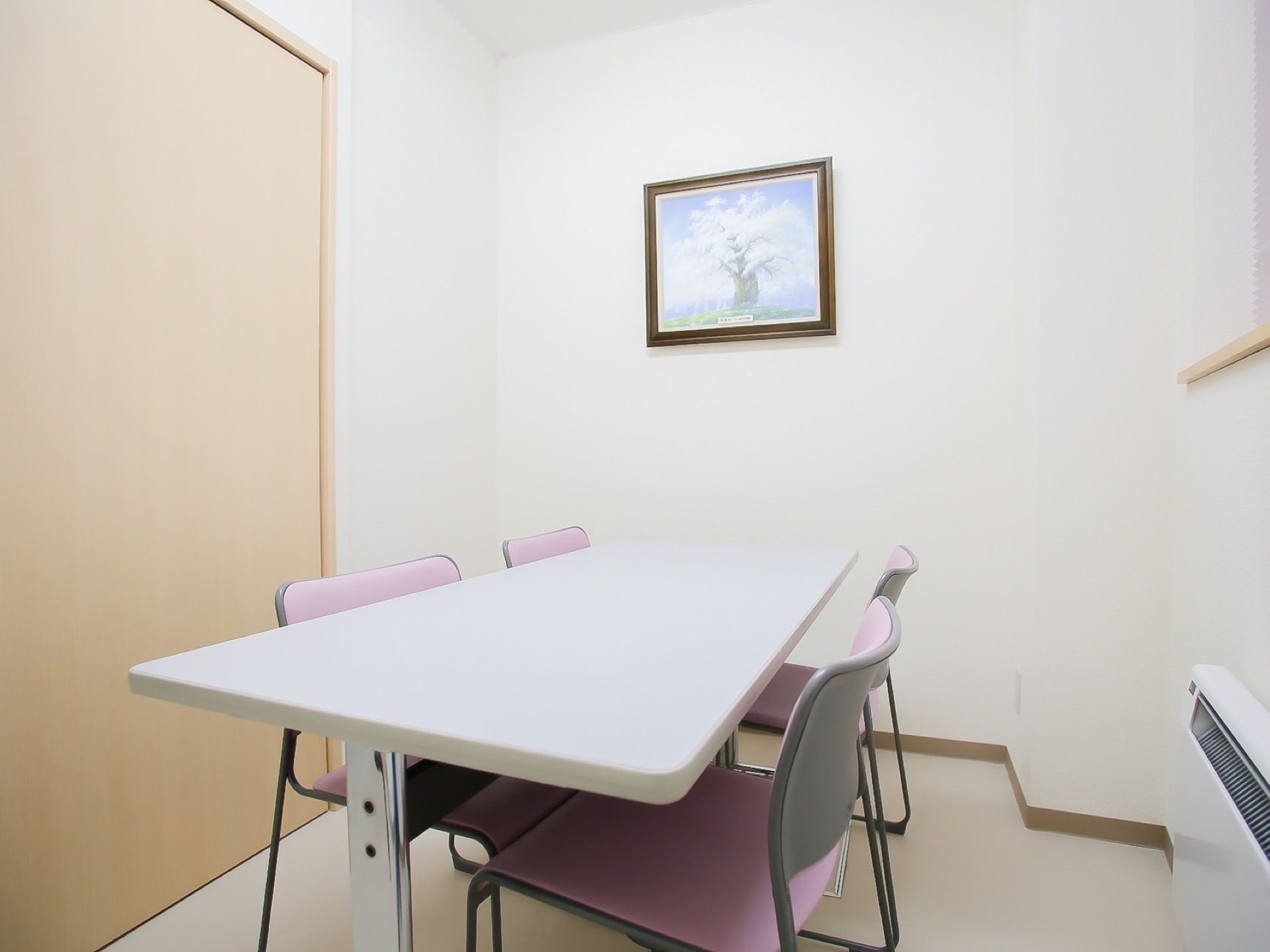 部屋の中央に白いテーブルと４脚のピンク色の椅子がある。壁に額縁に入った木の絵が飾られ、暖房器具が設置されている。