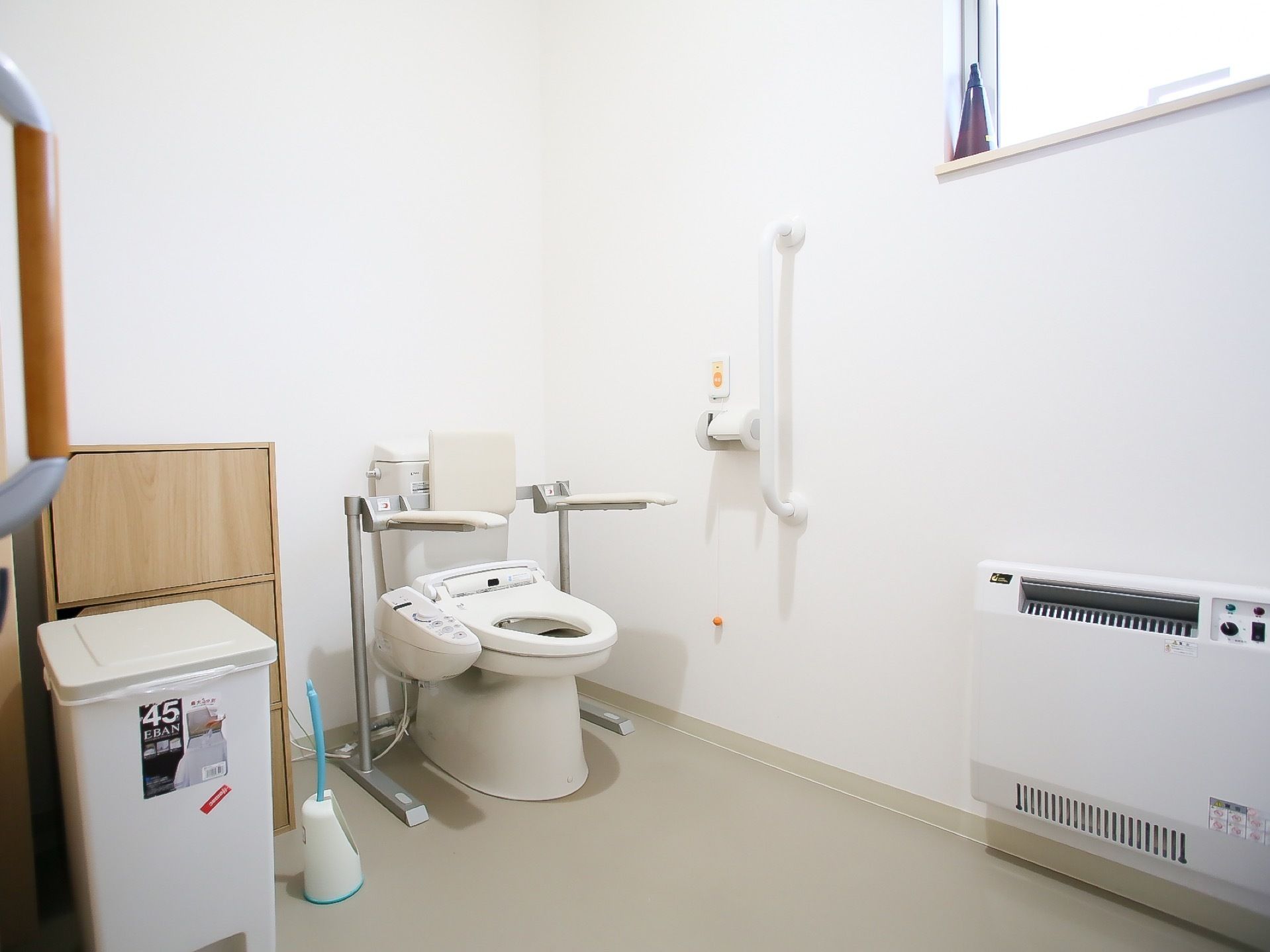 タンク付きの洋式トイレに手すりが設置されている。壁にナースコールと手すりと暖房器具が取りつけられている。