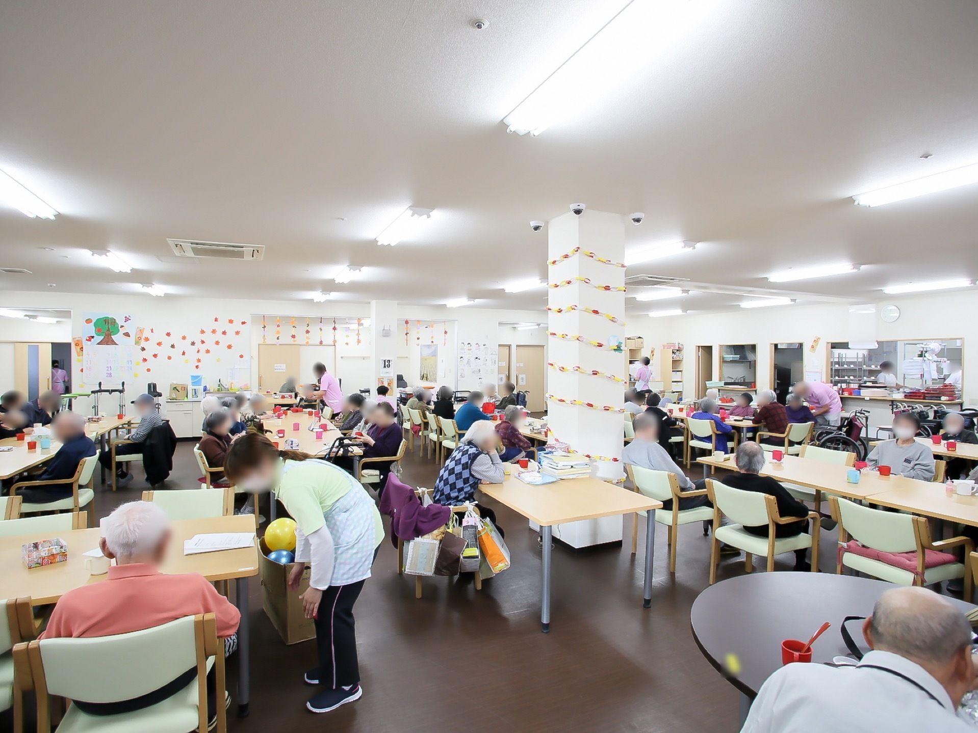 数本の構造柱が立っている大きな室内にたくさんの入居者様がいる。壁や柱に飾りつけがされ、天井に蛍光灯が設置されている。
