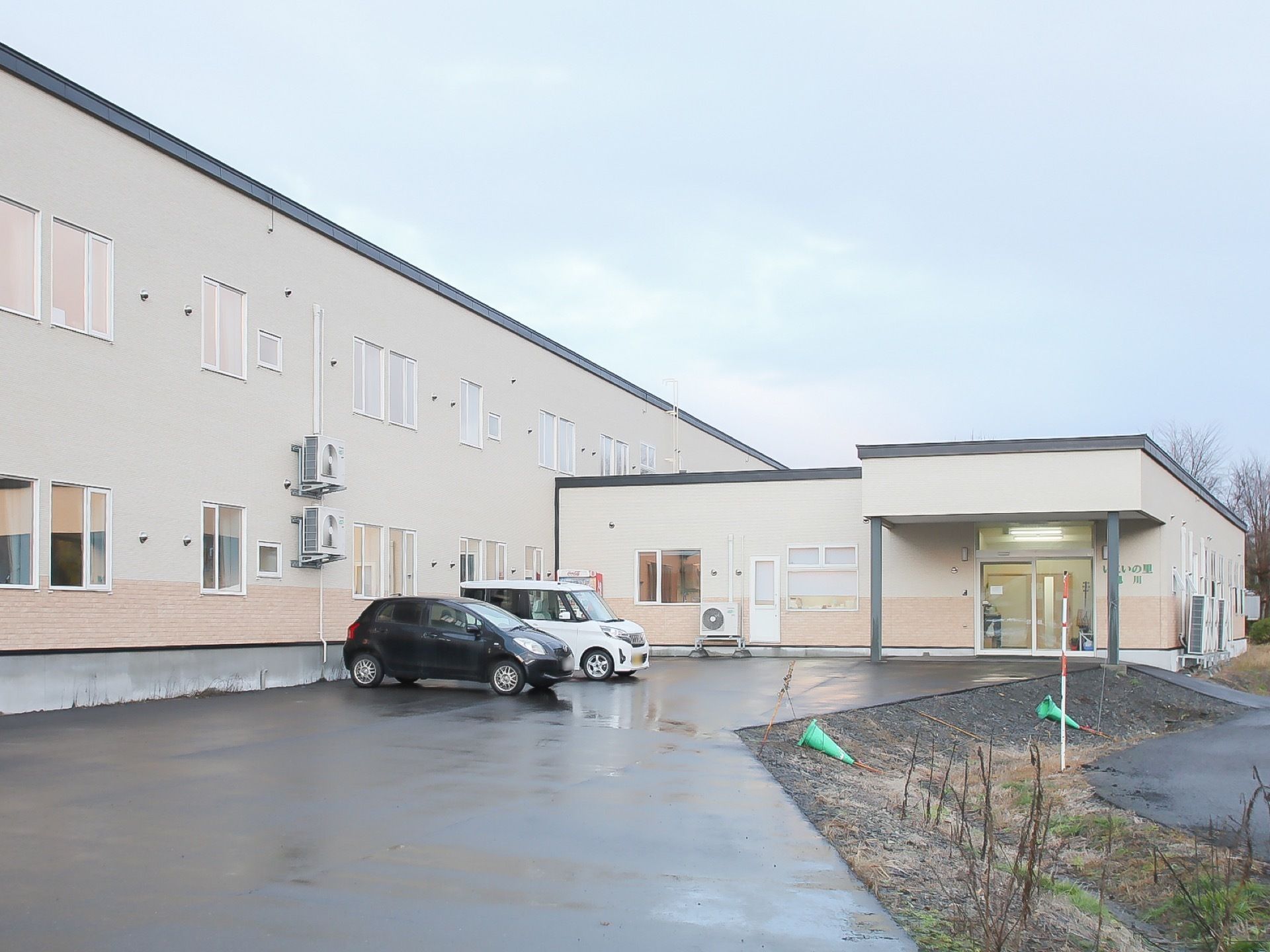 ２階建てのベージュ色の建物と１階建ての玄関が設置された建物がつながって建っている。アスファルトで整備された敷地の路面が濡れている。