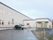 ２階建てのベージュ色の建物と１階建ての玄関が設置された建物がつながって建っている。アスファルトで整備された敷地の路面が濡れている。