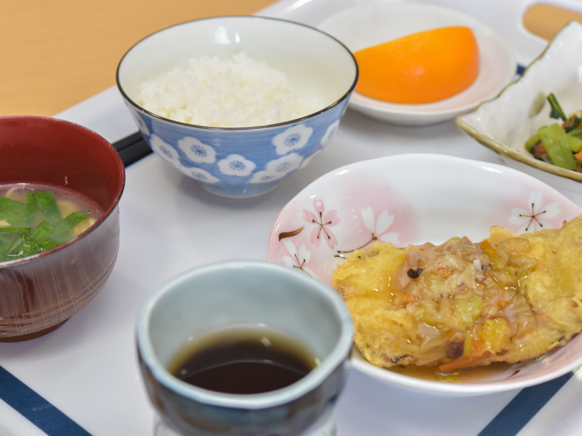 甘酢がかけられた魚のフライ、白ご飯、味噌汁、野菜の和え物が出されている。デザートにはオレンジがある。