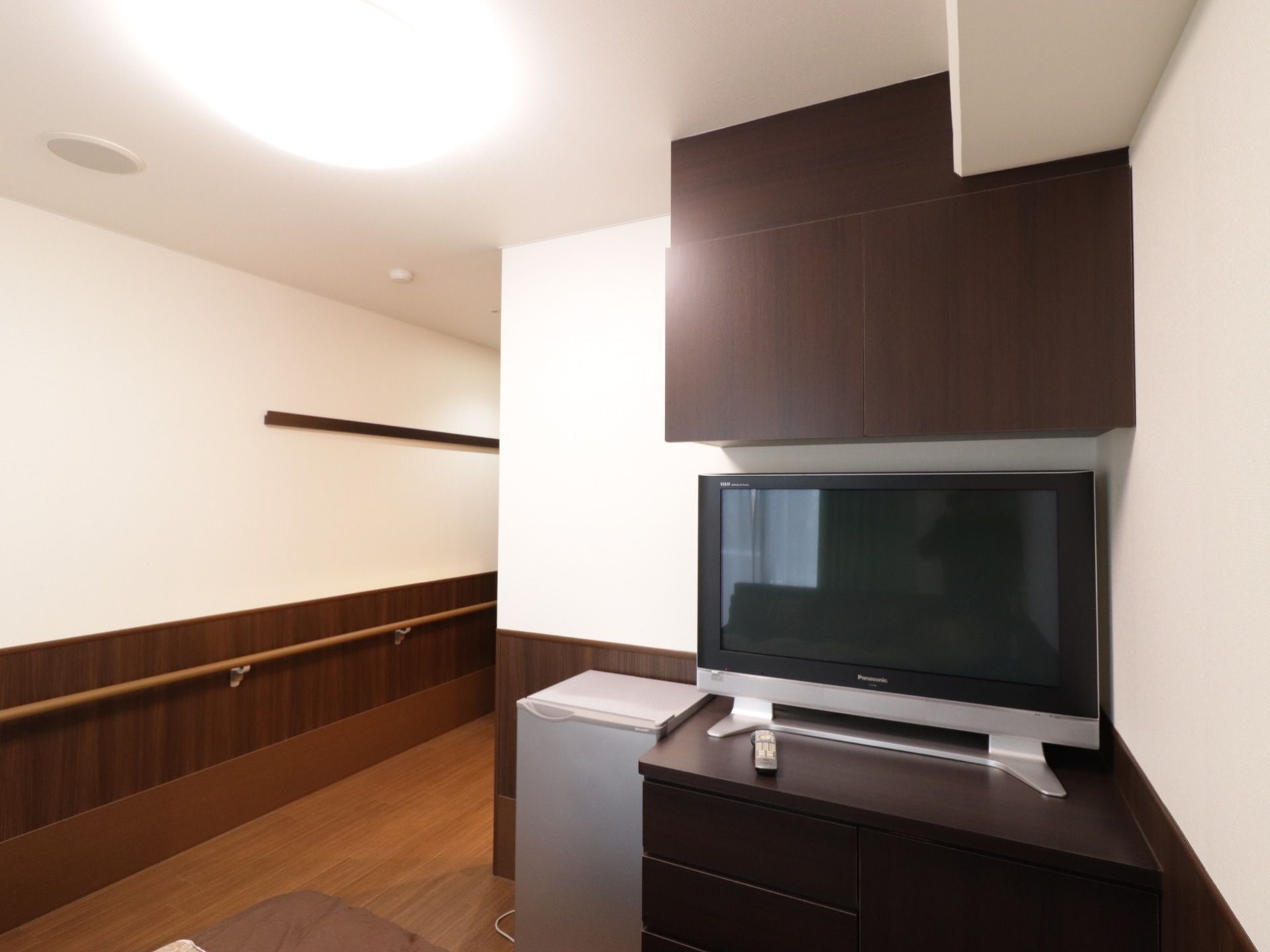 居室のベッドの足元に整理箪笥が置かれ、その上にはテレビがあり、さらに頭上には天井収納がある。機能性に富んだレイアウト。