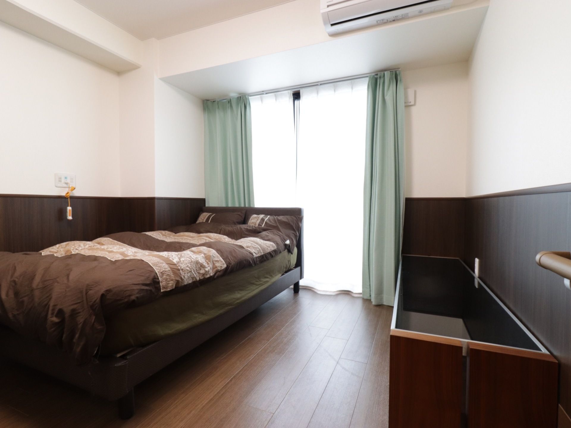 窓を正面に、ベッドを左手にとらえたアングルの居室。窓からの陽光で部屋が明るい。右手には背の低い横長の収納がある。