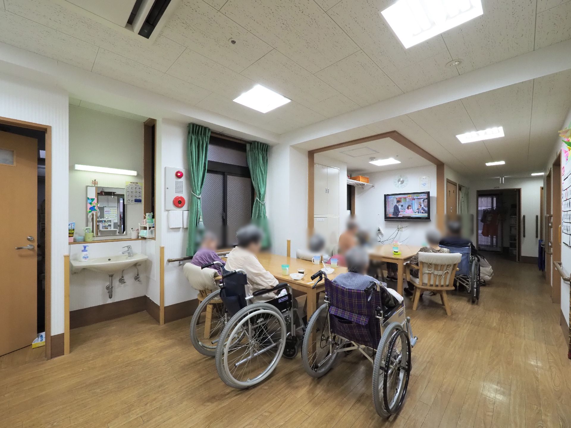 テーブルやイスが設置され、その周りで利用者が壁に掛かったテレビを観ている。一角には洗面台が設置されている。