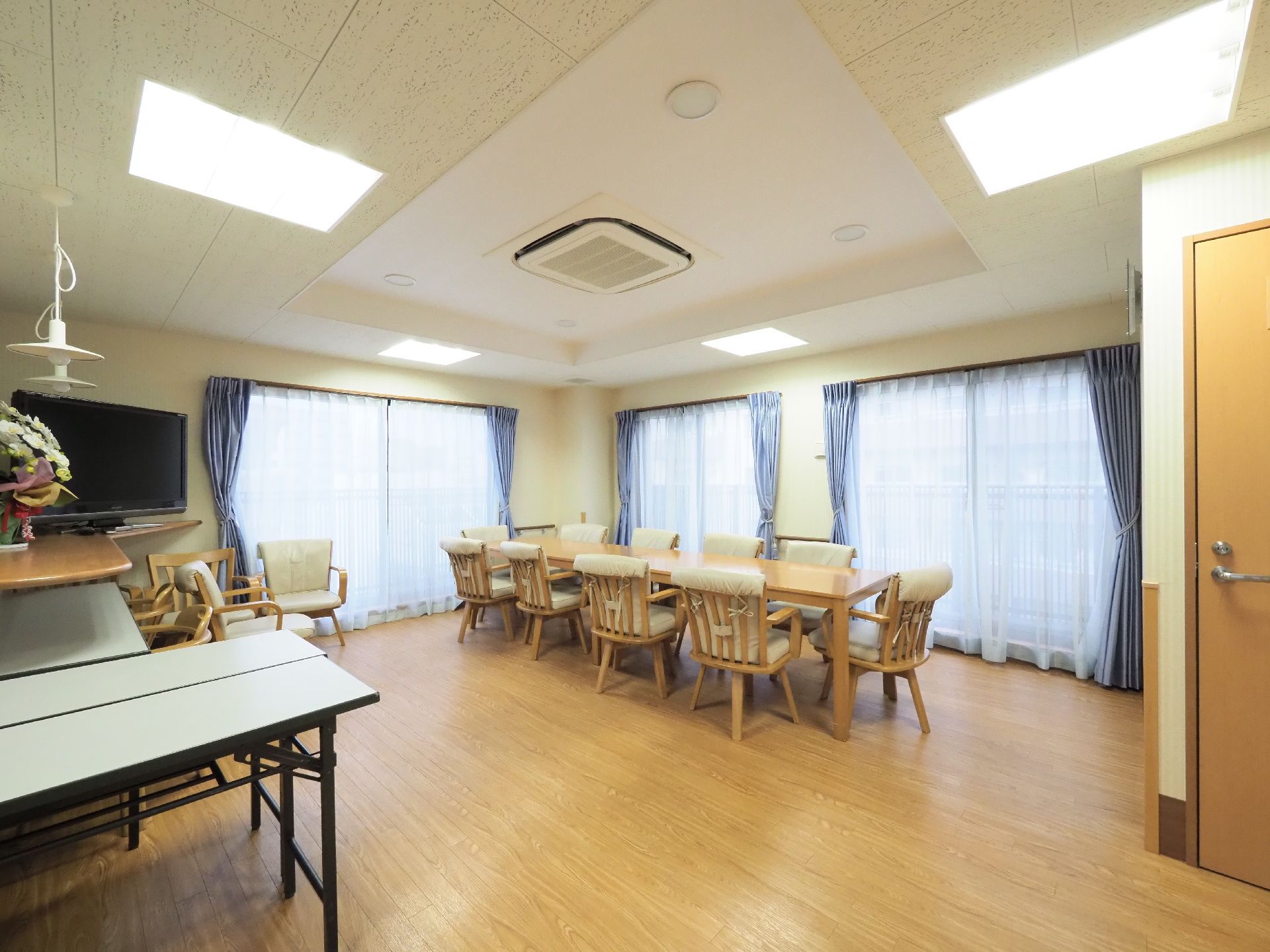 大きなガラスの窓が3か所ある。木製の机の周りには肘置きのついた椅子が10脚並べられている。少し離れた場所にも長机と椅子が見られる。