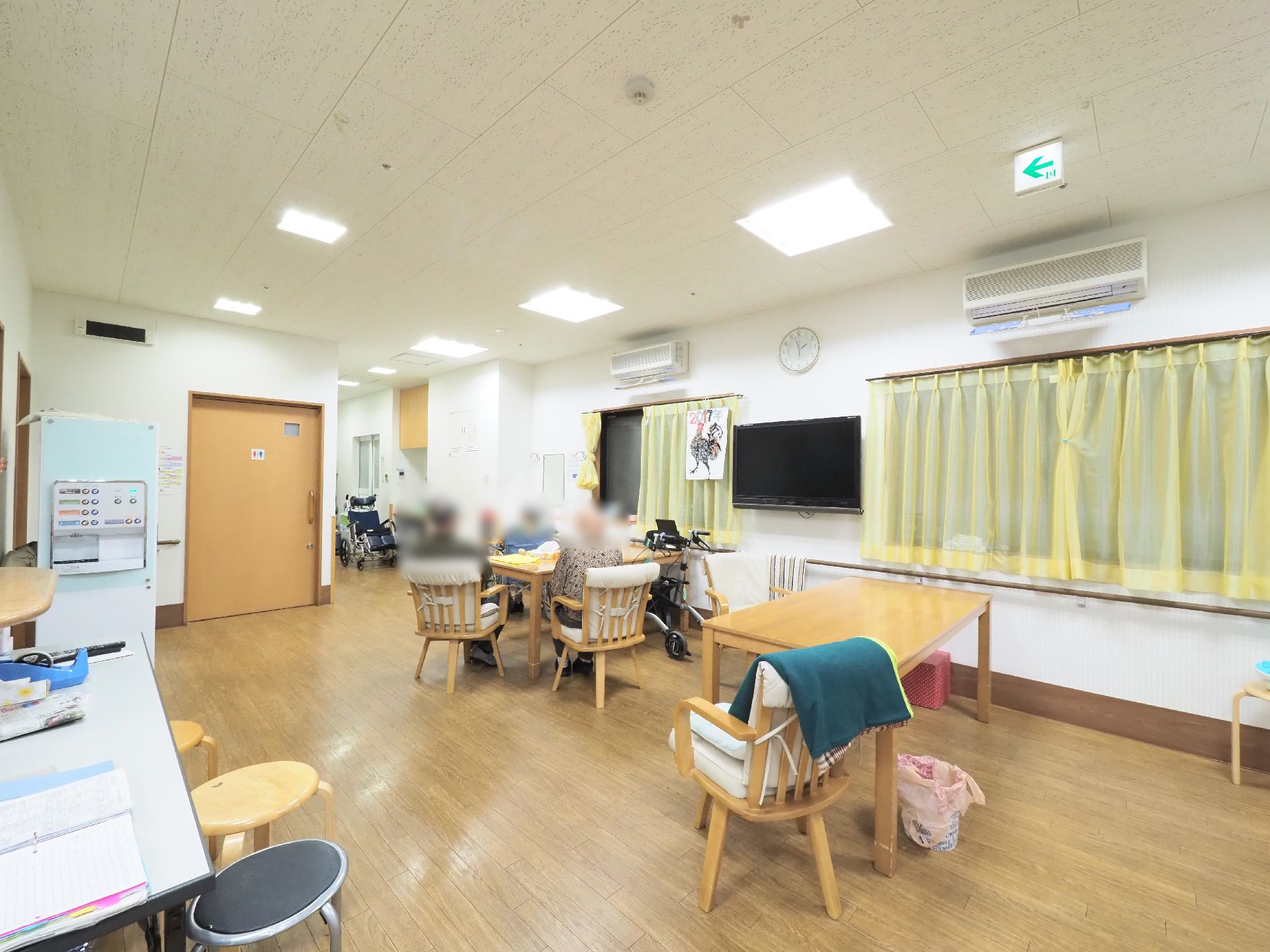 窓に薄い黄色のカーテンが取り付けられている、机と椅子があり、入居者たちが座っている正面には大きなテレビがある。