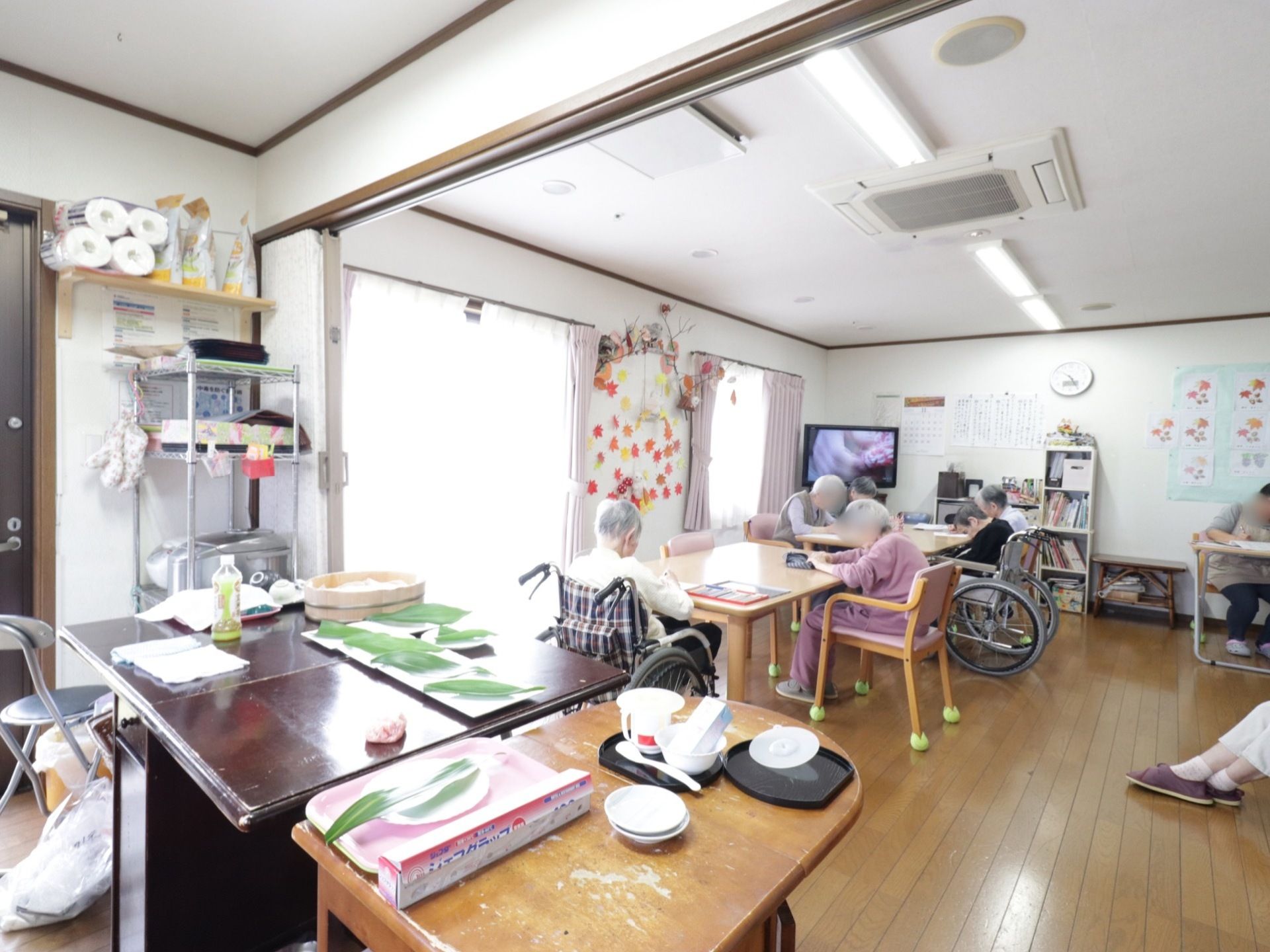 明るい光が差し込むスペースで、白い壁には手作りの飾りや入居者様の作品が飾られ温かみのある空間になっている。手前にキッチンが設置されている。