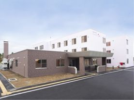 奥には白色の外観で清潔感のある居室の棟がある。各階には腰窓が設置されているため明るい居室になっている。
