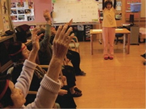開放的なスペースに入居者様が１列に並んで座っている。ホワイトボードの前に女性スタッフが立ち、一緒に体操をしている。
