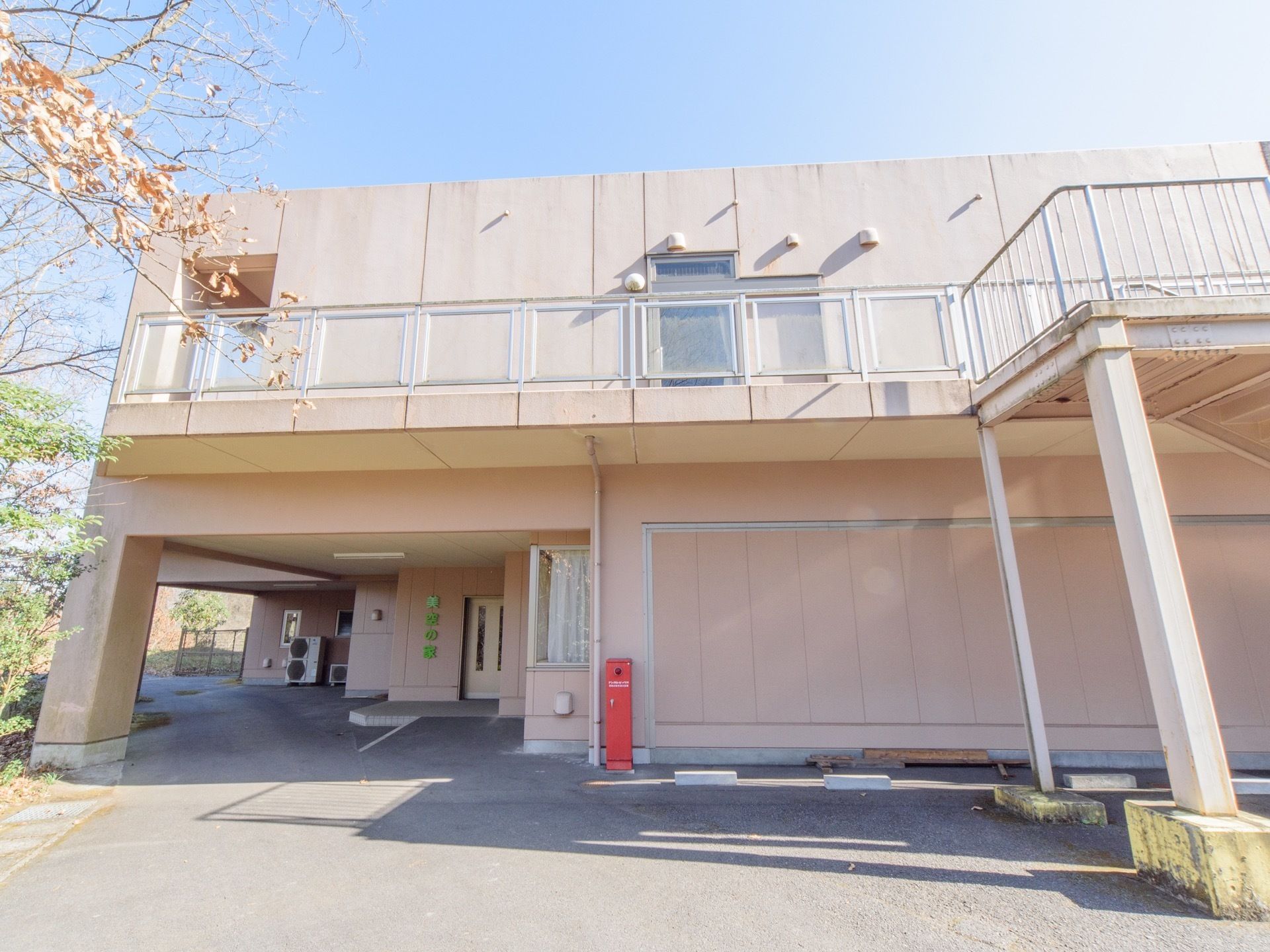 広々とした駐車場の奥に玄関がある。周辺に林があり自然が多い。２階建てのピンクベージュの建造物である。日当たりが良い。