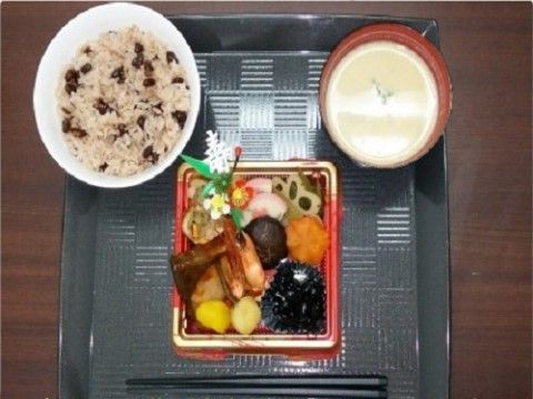 黒いモザイク模様のトレイにミニおせち、赤飯、汁椀が並べられている。おせちの内容は黒豆、栗きんとん、えび、魚、お煮しめなど。