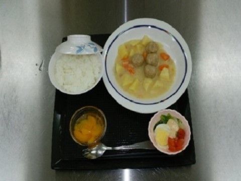 黒い正方形の盆に食事がのっている。時計回りにシチュー、サラダ、味噌汁、ご飯。手元にはスプーンが置かれている。