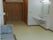 部屋の奥から出入り口を見た写真。ドアは引き戸になっている。左下にマットの敷かれていない電動ベッドがある。右の壁には洗面台が設置されている。