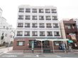 交差点の角に建つ５階建ての建物で、一階部分はレンガ風、二階以上は白色の外壁である。すぐ前にバス停がある。
