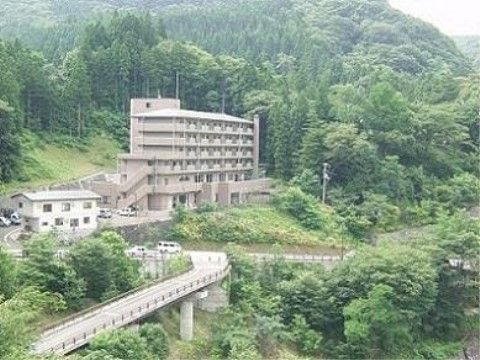 幹線道路が整備されている山あいに、6階建ての施設が建っている。施設には駐車スペースがあり、自然に囲まれている。