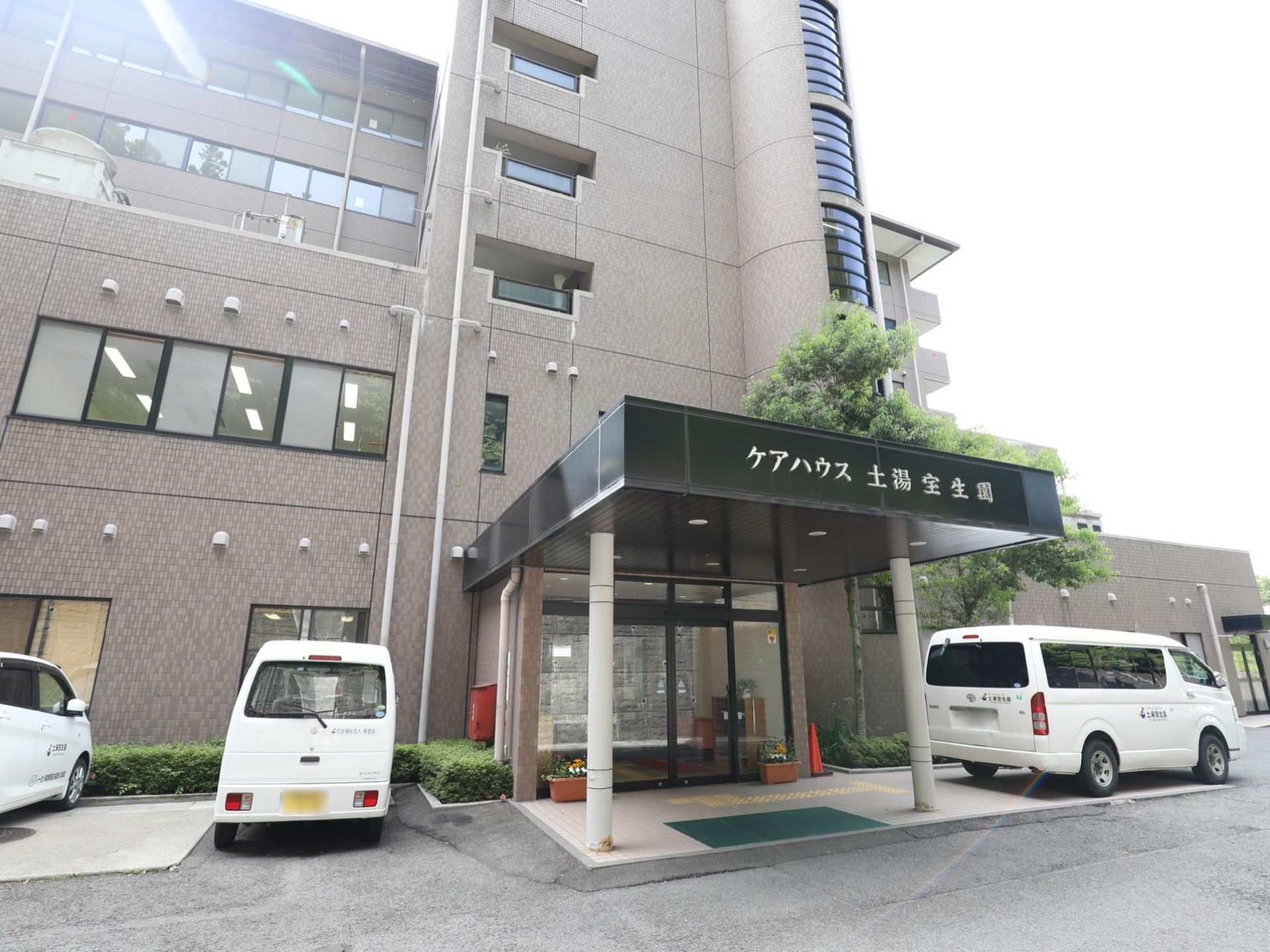 グレーの外観の建物である。玄関前には、どっしりとした屋根を設置している。建物の傍に低木の植栽を設けている。