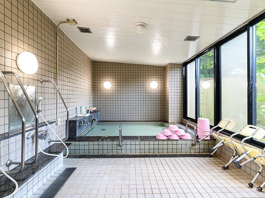 室内にある石張りの浴室である。天井まで高さのある窓際に大きな浴槽がある。洗い場にシャワーブースが並んでおり、縦向きの手すりがある。