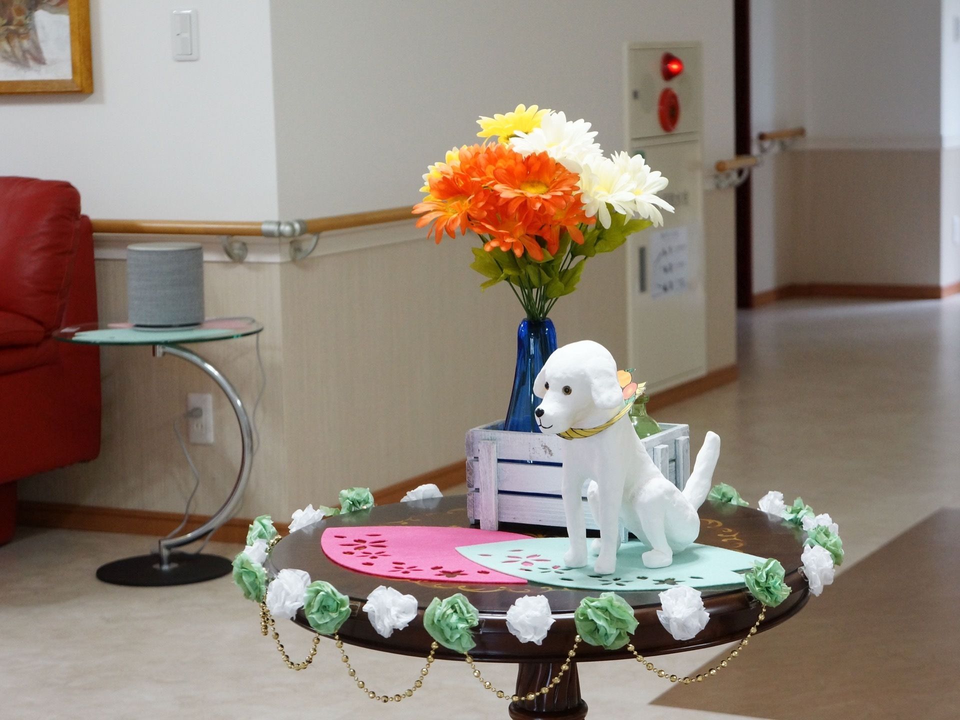 木製の丸いミニテーブルに、造花、白い犬の置物が飾られている。広々としたスペースの一角には、ソファや絵画が設置されている。