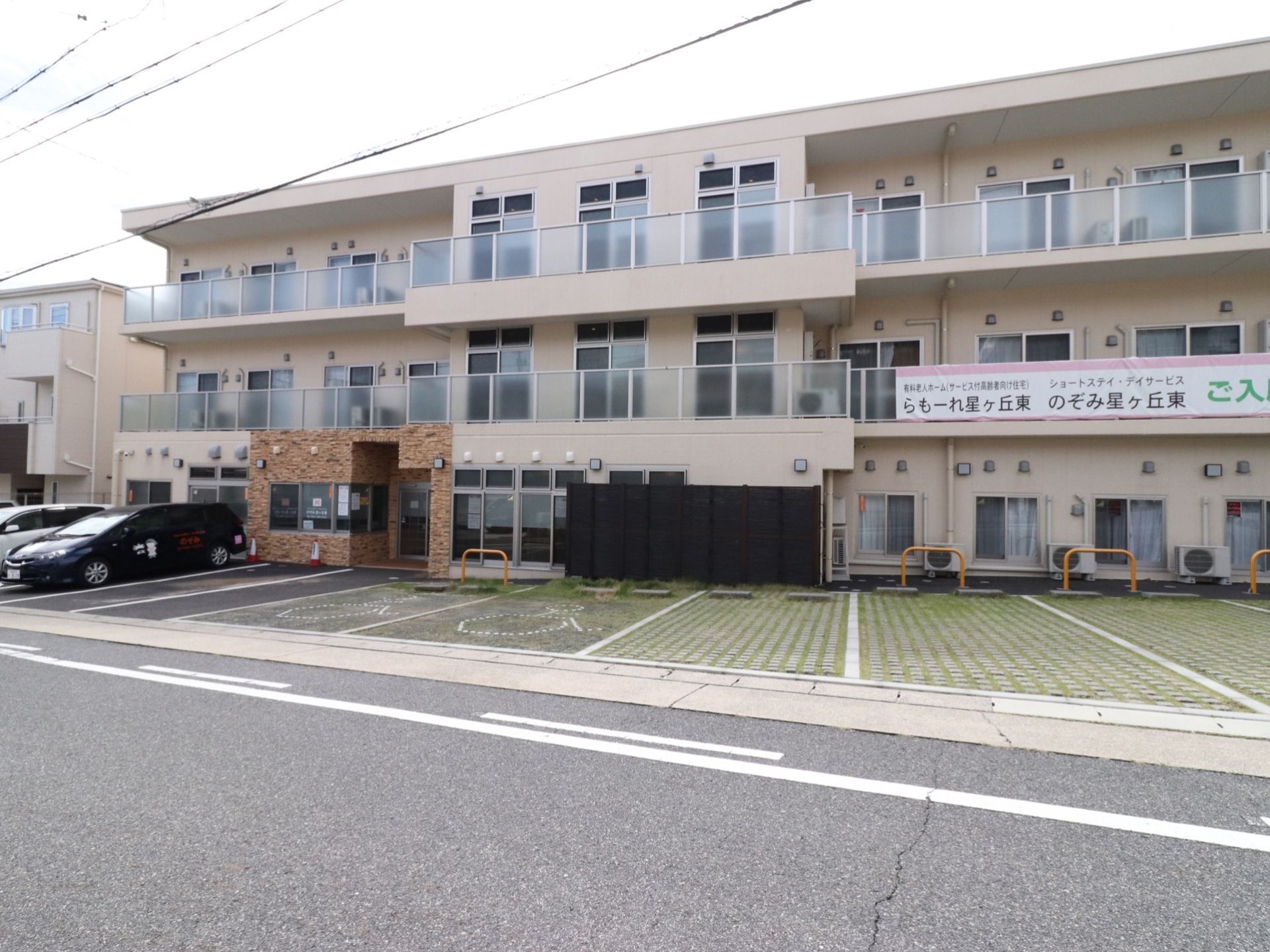 レンガ色とベージュのツートンカラーの建物である。3階建てで、大きな道路に面してベランダが設置してある。