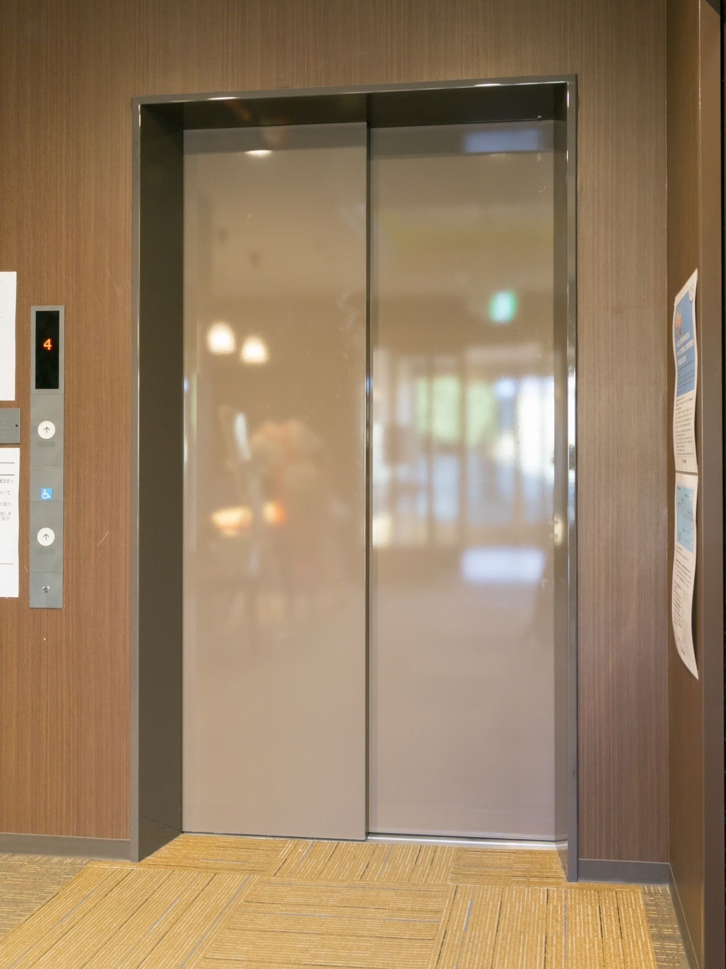 ベージュ色のエレベーターが1基設置されている。右側の壁にはチラシが2枚貼り付けられている。左側に操作ボタンがある。