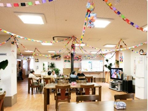 開放的な共有スペースで、天井にはカラフルな装飾がされている。入居者様がダイニングテーブルを囲んでテレビを楽しんでいる。