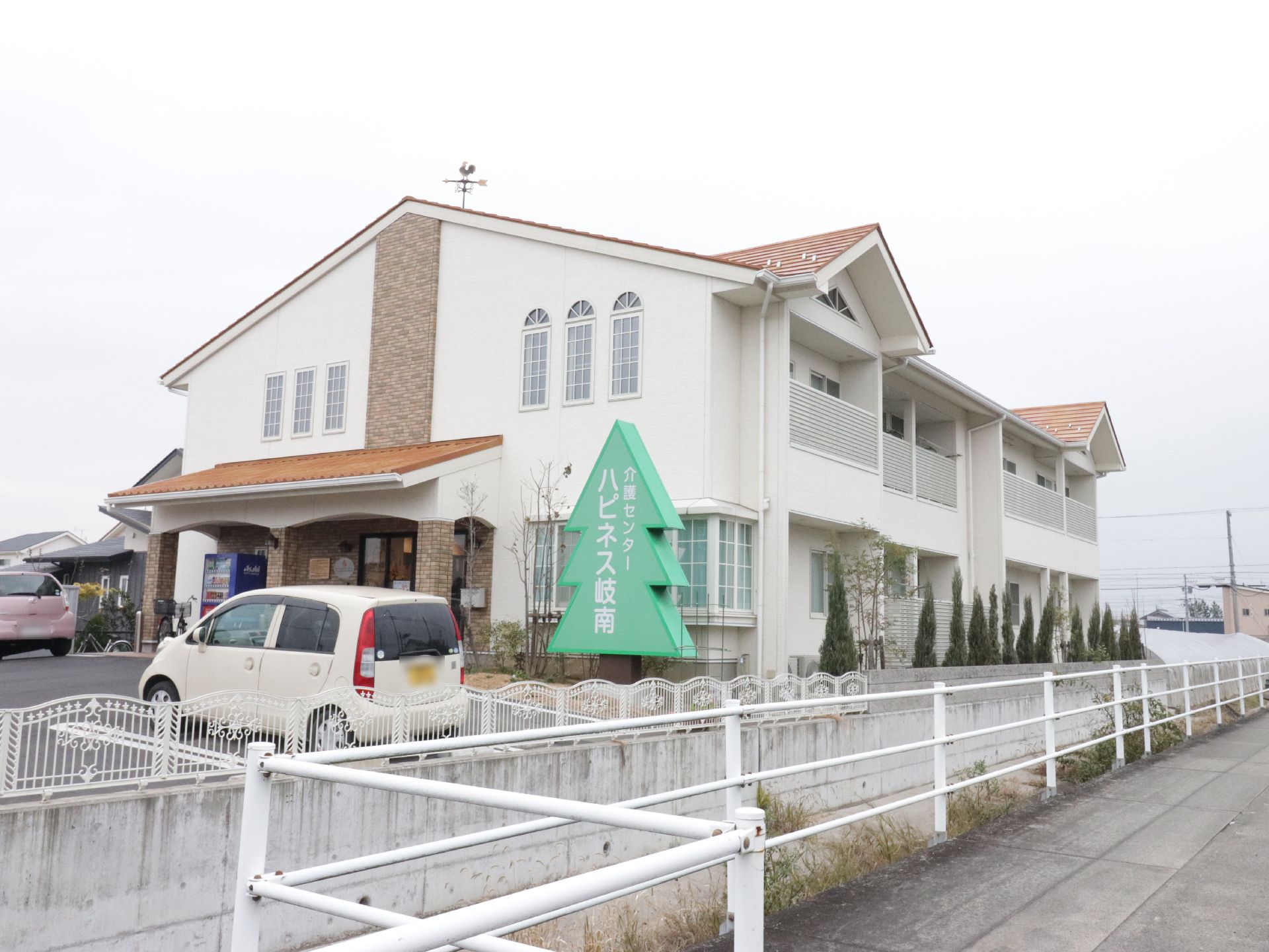 施設は2階建てで敷地面積が大きい。木を模った看板は大きく、施設名が書かれている。車が2台止められている。