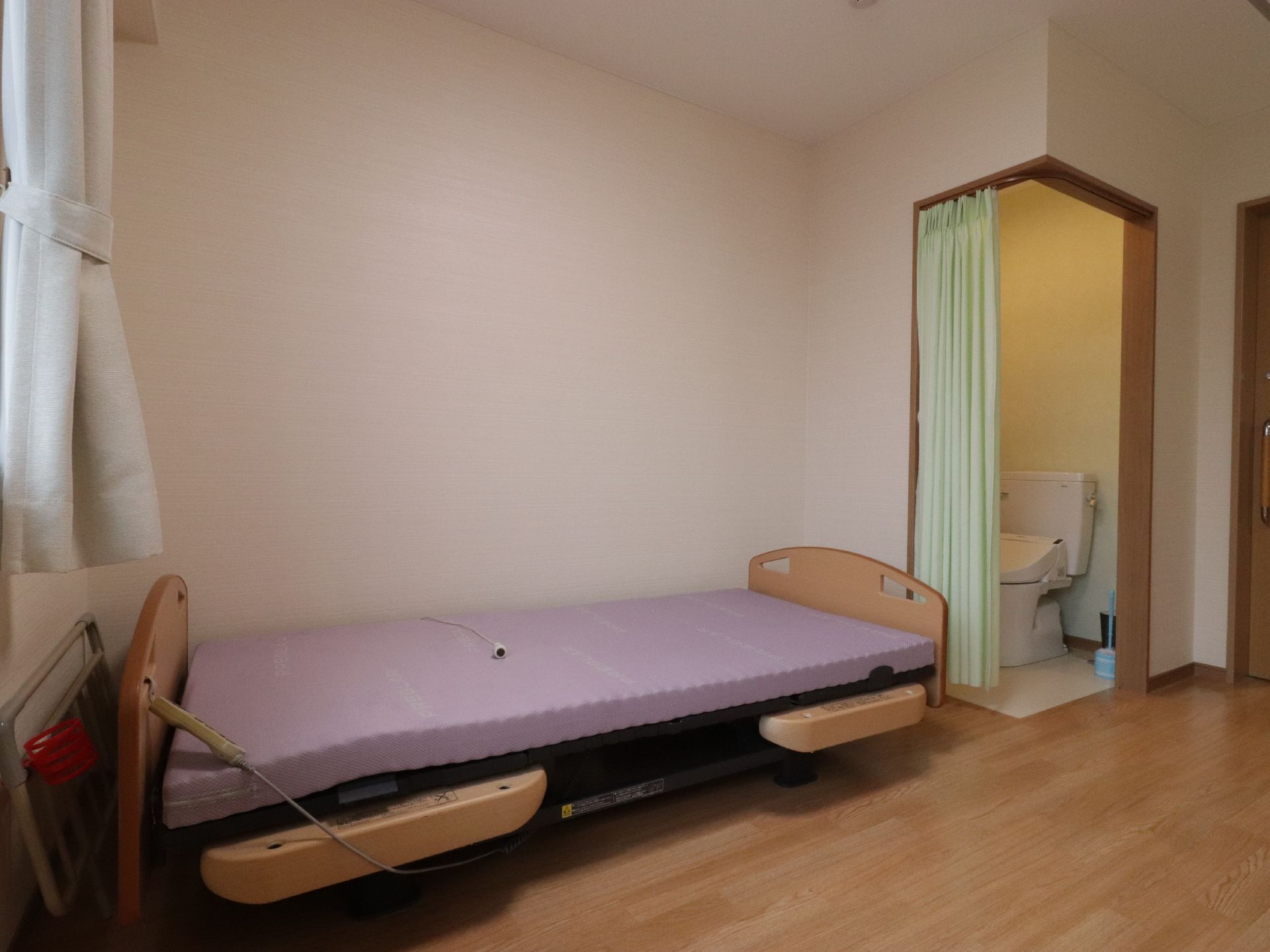 カーテンが掛かった近くにはベッドが置かれている。ベッドの足元側にはトイレが設置されている。トイレはカーテンで仕切られている。